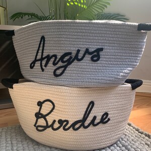 K&ouml;nnte beinhalten: Zwei graue und wei&szlig;e Korb-Aufbewahrungsk&ouml;rbe mit schwarzen Griffen. Der obere Korb hat den Namen "Angus" bestickt. Der untere Korb hat den Namen "Burdie" bestickt.