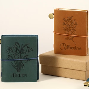 Puede incluir: Diarios de cuero en verde azulado y marrón claro con diseños florales y nombres. El diario verde azulado tiene el nombre "Helen" y el diario marrón claro tiene el nombre "Catherine". Los diarios se cierran con una banda elástica.