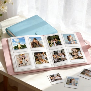 May include: A pink photo album with a light blue cover and a snap closure. The album is open, displaying several square photographs. A vase of flowers sits to the left. The album is on a white surface.