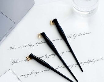 May include: Three black calligraphy pens with gold nibs and black handles. The pens are on a white surface with a handwritten note in black ink.