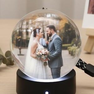 Può includere: Una palla di neve di vetro trasparente con una foto di una coppia in abito da sposa, posta su una base nera con un cordino bianco e nero. Sullo sfondo un terrario geometrico con un'orchidea rosa e luci fatate.