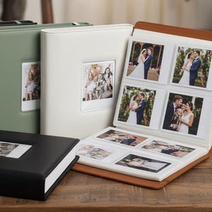 May include: A collection of photo albums in black, green, and white, with a brown album open to display photographs. The albums have a rectangular window on the front for inserting a photo. The open album displays several wedding photos.