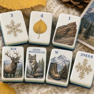 May include: Seven rectangular game tiles with rounded corners, featuring illustrations of a snowflake, an aspen leaf, a mountain landscape, an elk, a fox, a hiking boot, and a tree-like symbol, labeled with numbers, letters, and text.