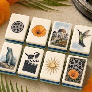 May include: A set of eight rectangular mahjong tiles with cream-coloured faces and blue-tinted sides. The tiles feature illustrations of a film reel, an orange poppy, the Griffith Observatory, a hummingbird, a seal, sunglasses, a clapperboard, and a sun.