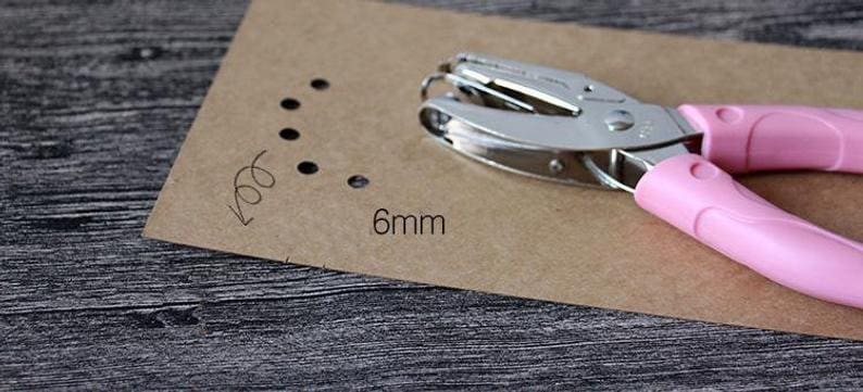 Puede incluir: Una perforadora plateada y rosa con una medida de 6 mm, que se muestra junto a un trozo de papel marr&oacute;n con agujeros perforados. Los mangos rosas de la perforadora tienen un dise&ntilde;o ergon&oacute;mico.