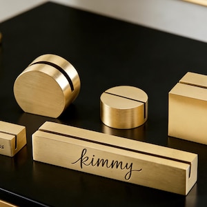 Op de afbeelding: Een verzameling goudkleurige metalen naambordhouders in verschillende vormen: een rechthoek, een kubus, een cilinder en een lange rechthoekige houder met de naam "kimmy" gegraveerd. De houder met het opschrift "Dress" is ook zichtbaar.