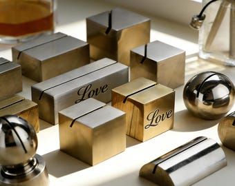 May include: A collection of metallic card holders in various shapes including cubes, spheres, and cylinders. Some pieces feature a brushed gold or silver finish, while two rectangular and cube-shaped holders are engraved with the word Love.
