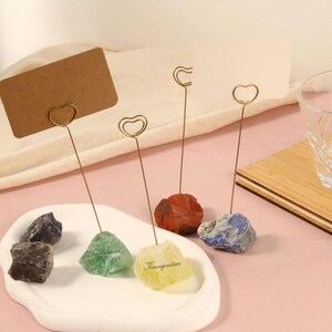 May include: A collection of place card holders featuring natural stone bases in various colors, including black, green, red, yellow, and blue. Each holder has a gold-colored wire with a heart-shaped top, holding a blank card. A clear glass and wooden tray are also present.