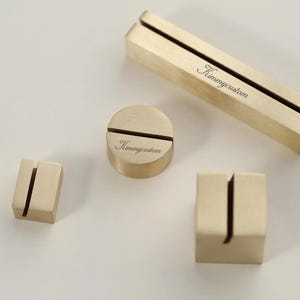 May include: A collection of brass table number holders in various shapes: rectangular, square, and circular. Each holder has a slot for a card. The text "Kimmycustom" is engraved on two of the holders.