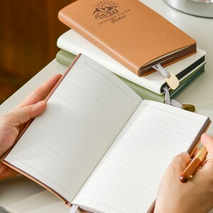 Op de afbeelding: Een bruin lederen dagboek met de tekst "Adventure Journal Richard" gegraveerd op de omslag. Het dagboek is open op een blanco pagina met gestreept papier. Een gouden bladwijzer is aan het dagboek bevestigd.