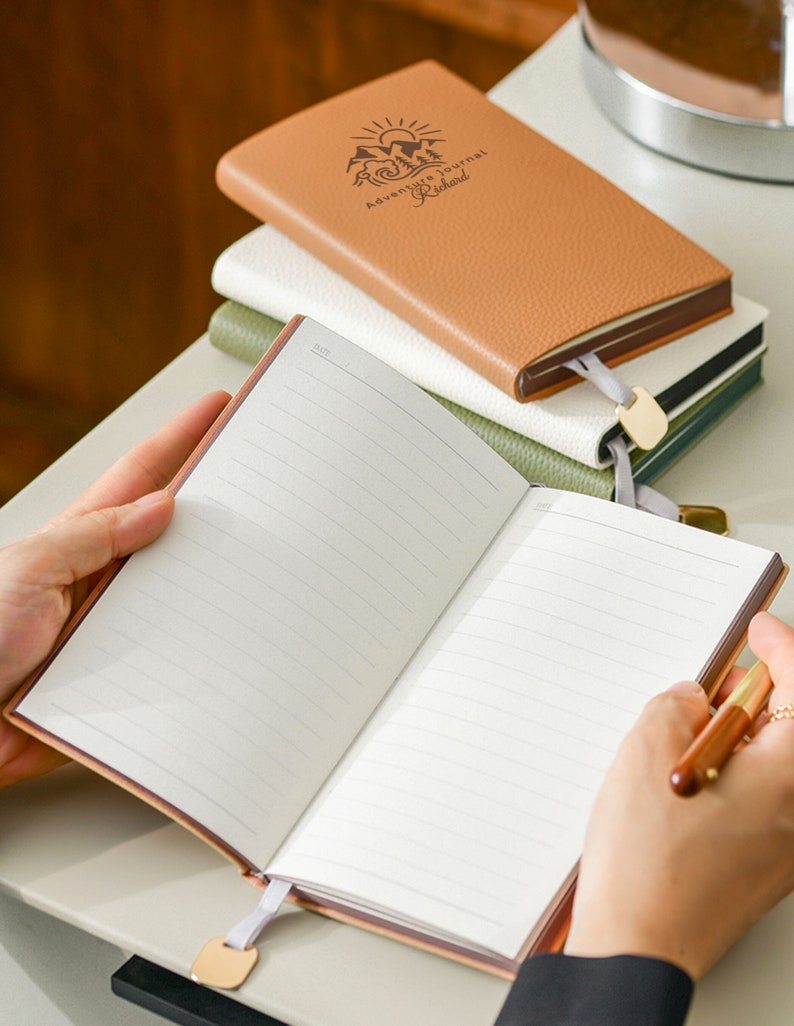 Op de afbeelding: Een bruin lederen dagboek met de tekst "Adventure Journal Richard" gegraveerd op de omslag. Het dagboek is open op een blanco pagina met gestreept papier. Een gouden bladwijzer is aan het dagboek bevestigd.