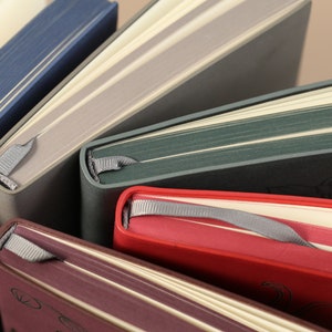 May include: Five different colored notebooks with ribbon bookmarks. The notebooks are stacked on top of each other. The top notebook is blue, the second is gray, the third is green, the fourth is red, and the bottom is burgundy.