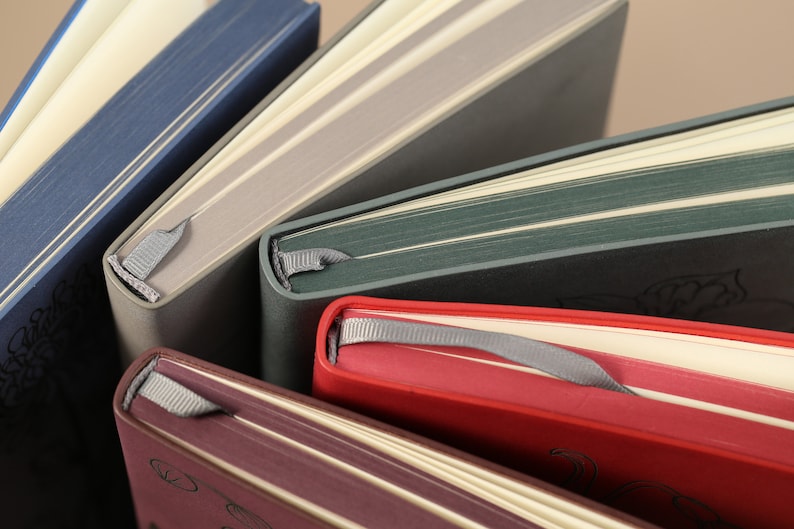 May include: Five different colored notebooks with ribbon bookmarks. The notebooks are stacked on top of each other. The top notebook is blue, the second is gray, the third is green, the fourth is red, and the bottom is burgundy.
