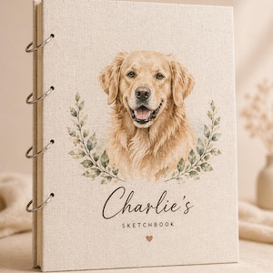 Puede incluir: Un cuaderno de bocetos beige con encuadernación de anillas metálicas. La portada presenta un retrato en acuarela de un perro Golden Retriever, rodeado de hojas verdes. El nombre "Charlie's" está escrito en cursiva, con "SKETCHBOOK" debajo.