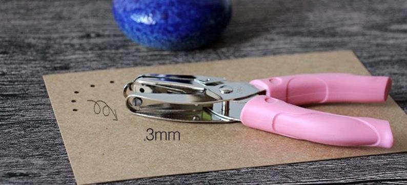 Puede incluir: Perforadora rosa y plateada con una marca de medici&oacute;n de 3 mm en papel marr&oacute;n. La herramienta tiene mangos rosas y est&aacute; dise&ntilde;ada para crear peque&ntilde;os agujeros en papel. Un jarr&oacute;n de cer&aacute;mica azul est&aacute; en el fondo.
