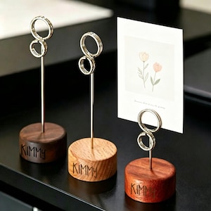 May include: Three circular wooden bases in varying brown tones, each featuring the name KIMMY engraved on the side. Each base supports a tall, thin silver metal rod topped with a double-ring clip, one holding a white card with a floral illustration.