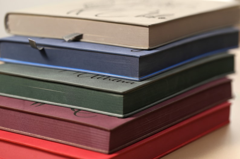 May include: A stack of five hardcover books with different colored covers. The top book is beige, the second is blue, the third is gray, the fourth is burgundy, and the bottom book is red.