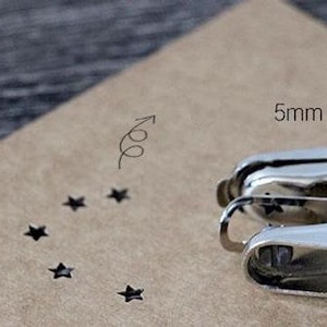 Puede incluir: Una perforadora de metal plateado junto a un trozo de papel marr&oacute;n con recortes en forma de estrella. El papel tiene una medida de 5 mm y un gr&aacute;fico de flecha. La perforadora est&aacute; enfocada, lo que sugiere un tema de manualidades o papeler&iacute;a.