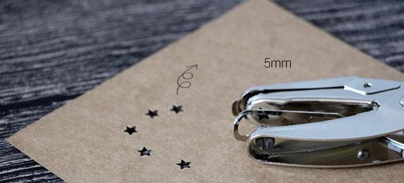 Puede incluir: Una perforadora de metal plateado junto a un trozo de papel marr&oacute;n con recortes en forma de estrella. El papel tiene una medida de 5 mm y un gr&aacute;fico de flecha. La perforadora est&aacute; enfocada, lo que sugiere un tema de manualidades o papeler&iacute;a.