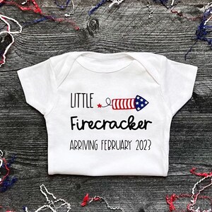 May include: White baby onesie with a red, white and blue firework graphic and the text "Little Firecracker Arriving February 2023?"