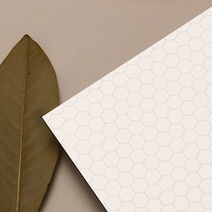 Puede incluir: Una taza de cerámica rosa llena de té marrón se encuentra sobre una superficie beige junto a una hoja marrón seca. Una hoja de papel blanca con un patrón hexagonal es parcialmente visible en el fondo.