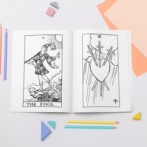 May include: A black and white coloring book featuring illustrations of tarot cards. The book is open to two pages, showing the "The Fool" card and the "Three of Swords" card. The book is surrounded by colored pencils and small geometric shapes.