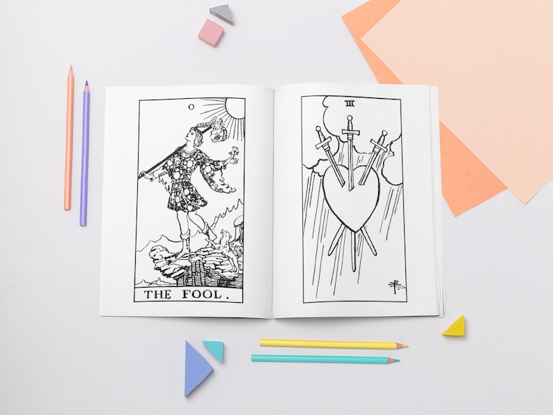 May include: A black and white coloring book featuring illustrations of tarot cards. The book is open to two pages, showing the "The Fool" card and the "Three of Swords" card. The book is surrounded by colored pencils and small geometric shapes.