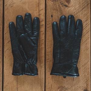 May include: A pair of black leather gloves with a textured surface. The gloves are laid flat on a wooden surface.
