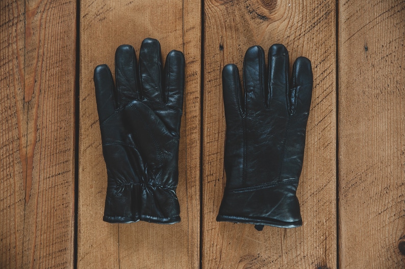 May include: A pair of black leather gloves with a textured surface. The gloves are laid flat on a wooden surface.
