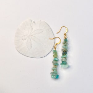 Puede incluir: Pendientes colgantes con ganchos dorados. Cada pendiente presenta una cadena vertical de pequeñas piedras irregulares, de color azul claro y verde. Un dólar de arena blanco en el fondo.