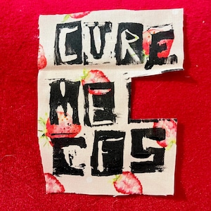 May include: A fabric patch with the words "CURE ME EFS" printed in black block letters. The fabric has a strawberry pattern on a white background. The patch is set against a red background.