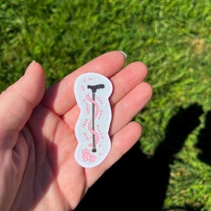 Op de afbeelding: Een gestanste sticker met een zwarte wandelstok omwikkeld met een roze lint, tegen een lichtblauwe achtergrond met kleine roze kersenbloesems en de tekst "your cane doesn't ruin your outfit" geschreven in roze cursief schrift.