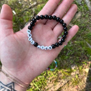 May include: A stretch bracelet composed of smooth, round, glossy black beads interspersed with white circular beads featuring the black printed letters that spell out the words FALL RISK. The bracelet is circular and features a uniform bead size throughout.