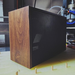 May include: A rectangular computer case with a wood grain finish on one side and a black perforated metal front panel. The case is sitting on a light-colored surface with small wooden pegs around it. The case is likely for a desktop computer.