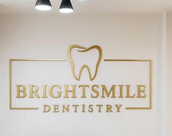 May include: A gold-colored sign on a white wall for Brightsmile Dentistry. The sign features the words "BRIGHTSMILE" above "DENTISTRY" in a gold, sans-serif font. A gold tooth graphic is above the text. The sign is framed by a gold rectangle.