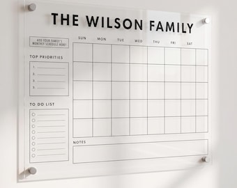 May include: A clear acrylic wall calendar featuring the text THE WILSON FAMILY at the top. It includes a weekly grid, a section for top priorities, a to-do list with checkboxes, and a notes area, all printed in black with silver hardware.