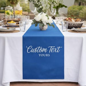 May include: A white tablecloth is set for a meal, with a vibrant blue table runner. The runner has the words "Custom Text Yours" in white script. The table is set with glasses, plates, and floral centerpieces.