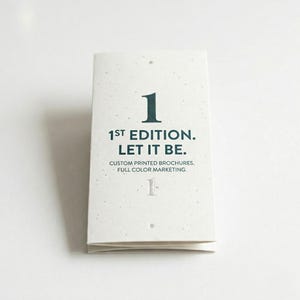 May include: A folded brochure with the text "1", "1ST EDITION. LET IT BE.", "CUSTOM PRINTED BROCHURES. FULL COLOR MARKETING." The brochure is white with a speckled texture and dark teal lettering.