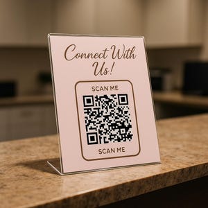 May include: A clear acrylic sign with a light pink background. The sign reads "Connect With Us!" in a script font. Below is a QR code with "SCAN ME" above and below it. The sign is on a brown surface.
