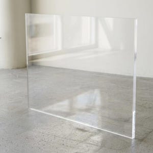 May include: A large, clear acrylic panel stands upright on a concrete floor. The transparent sheet reflects the surrounding environment, including a window and a potted plant. The panel's edges are clean and sharp.