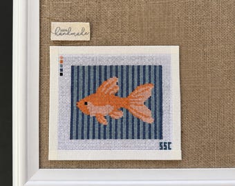Op de afbeelding: Een naaldpunt canvas met een oranje goudvis gecentreerd tegen een achtergrond van verticale donkerblauwe strepen. Het canvas bevat links een kleine kleurenpaletgids en in de rechterbenedenhoek de letters SSC.