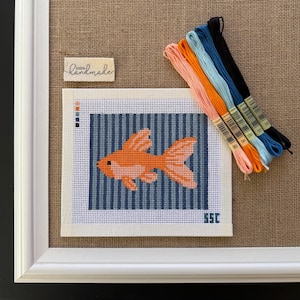 May include: A framed needlepoint featuring an orange goldfish against a blue striped background. The artwork is accompanied by colorful embroidery floss and a small tag that reads "handmade". The piece is set against a burlap background.