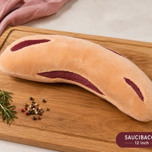 May include: A plush sausage-shaped toy, approximately 30 cm long, in tan and burgundy, on a wooden cutting board. The toy has burgundy accents. Rosemary sprigs and peppercorns are scattered nearby. The text "SAUCIBACON" is visible.