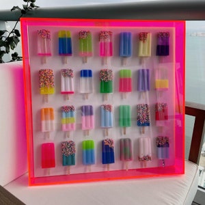 Puede incluir: Un marco rosa ne&oacute;n con fondo blanco muestra una colecci&oacute;n de 24 paletas de hielo coloridas con chispas. Cada paleta de hielo es de un color diferente y tiene un patr&oacute;n de chispas diferente.