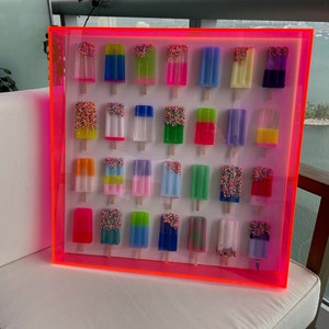 Puede incluir: Una pieza de arte de pared enmarcada de color rosa ne&oacute;n con 24 paletas de colores con chispas. Las paletas est&aacute;n dispuestas en cuatro filas de seis.