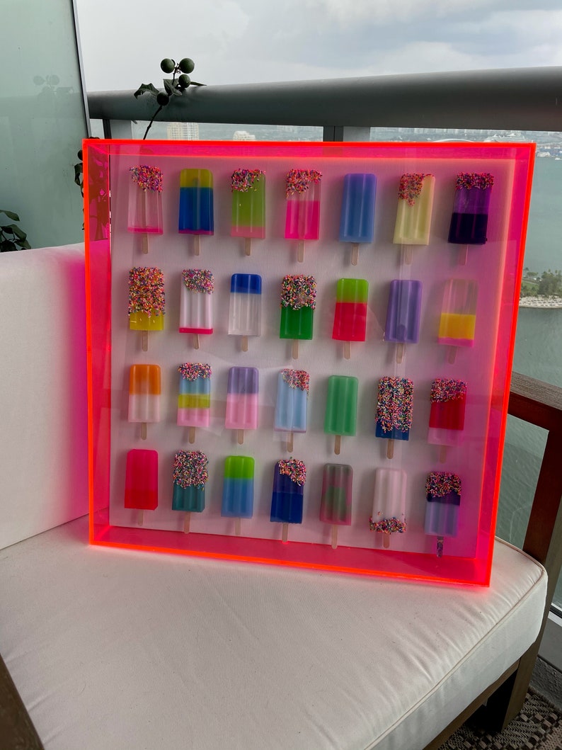 Puede incluir: Una pieza de arte de pared enmarcada de color rosa ne&oacute;n con 24 paletas de colores con chispas. Las paletas est&aacute;n dispuestas en cuatro filas de seis.