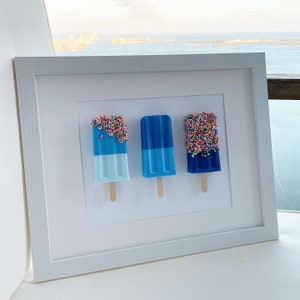 Könnte beinhalten: Drei blaue Eis am Stiel mit weißen und blauen Streifen und bunten Streuseln oben sind in einem weißen Rahmen gerahmt.