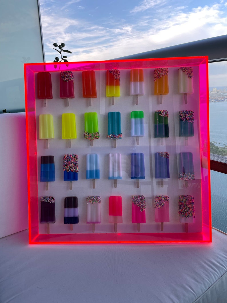 Puede incluir: Una pantalla enmarcada de 20 paletas de hielo coloridas con chispas. Las paletas de hielo est&aacute;n dispuestas en cuatro filas de cinco, con cada fila presentando un color diferente. El marco es rosa y tiene un borde rosa ne&oacute;n.