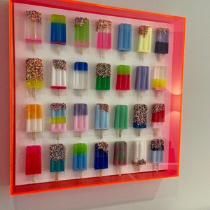 Puede incluir: Una pieza de arte de pared enmarcada que presenta 25 esculturas coloridas con forma de paleta de helado con chispas. Las paletas de helado est&aacute;n dispuestas en cinco filas de cinco, con cada paleta de helado teniendo un color y un dise&ntilde;o diferente. El marco es de color naranja brillante.