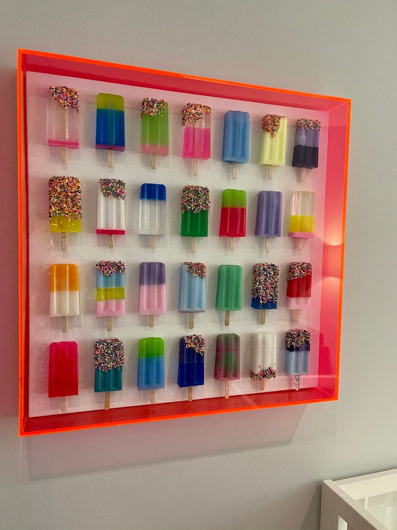 Puede incluir: Una pieza de arte de pared enmarcada que presenta 25 esculturas coloridas con forma de paleta de helado con chispas. Las paletas de helado est&aacute;n dispuestas en cinco filas de cinco, con cada paleta de helado teniendo un color y un dise&ntilde;o diferente. El marco es de color naranja brillante.